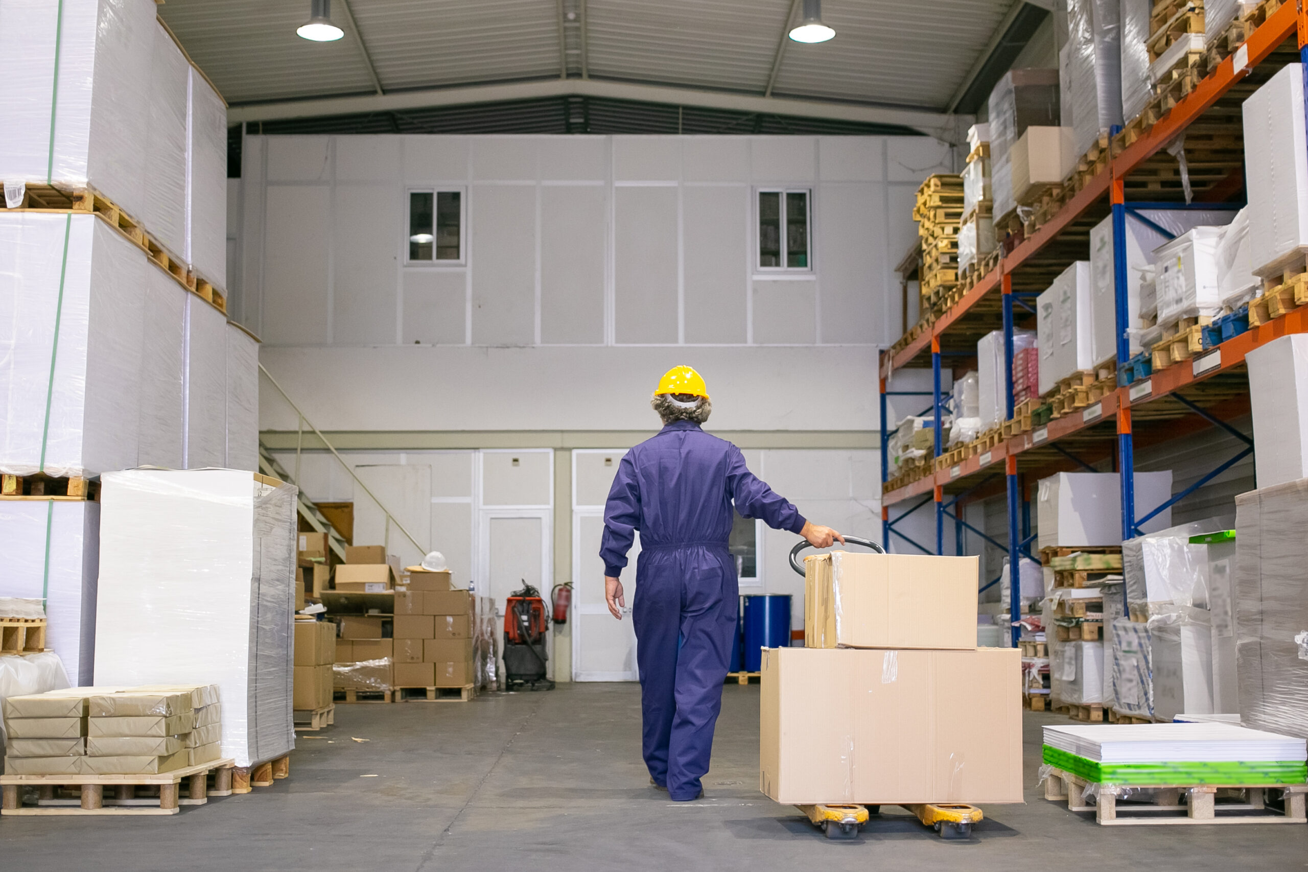 senior logistic worker hardhat uniform walking warehouse wheeling palette jack back view full length labor logistics concept 1 scaled